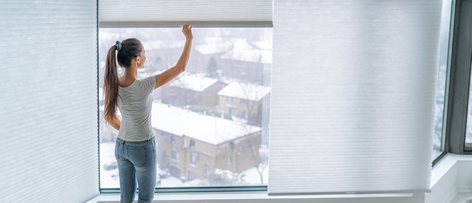 a woman opening her pleated blinds overlooking a snowy suburban area
